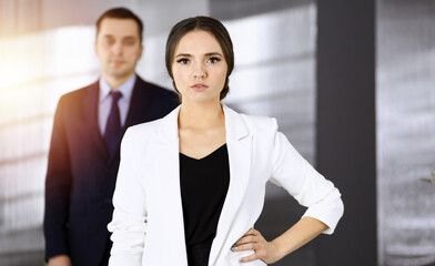 Young intelligent businesswoman is standing straight in a sunny modern office with her colleague at the background. Concept of successful business