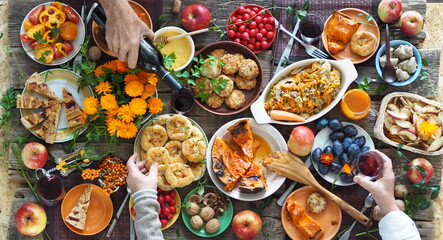 Country style. Thanksgiving table. Lots of food. Guests pour red wine into a glass. Guests are holding food with their hands over the set table. View from above.