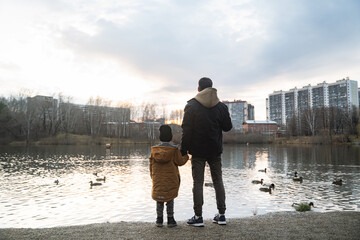 Silhouettes of a father and son standing by the lake, ducks are swimming, the sun is setting, against the background of multi-storey buildings, one parent is walking with a child in the park
