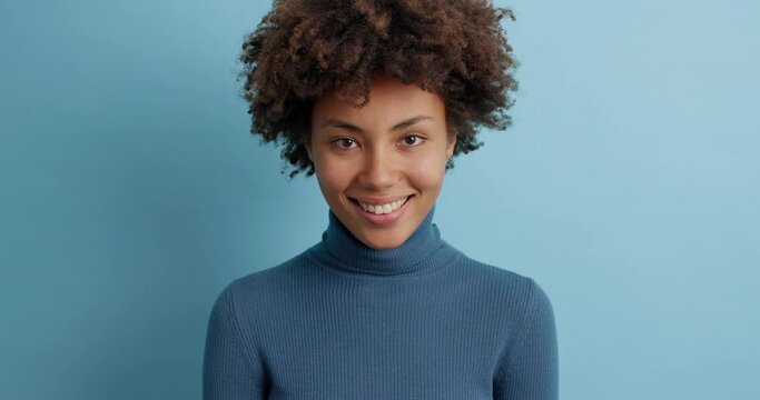 Glad smiling young Afro American woman with perfect white teeth has positive expression looks happily at camera dressed in blue poloneck chuckles as hears something good or funny poses indoor.