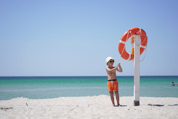Naklejka na ściany i meble child in a Panama hat and sunglasses rests on the beach, holding an orange lifebuoy.
