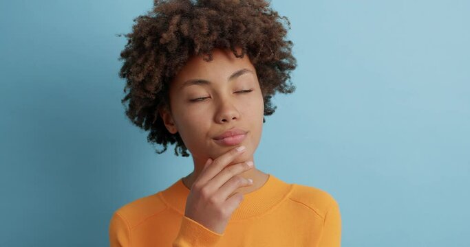 Thoughtful young Afro American woman looks somewhere pensively thinks deeply about something tries to make decision in mind wears casual orange jumper poses against blue background. Let me think