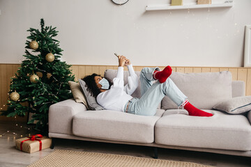 Young african american wearing protective mask, typing a message while lying on her sofa in Christmas-decorated room. Social distance during Covid-19 pandemic concept