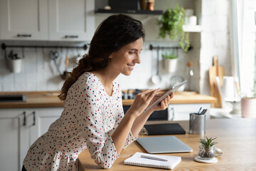 Happy young Caucasian woman at home kitchen use modern tablet communicate online. Smiling female browse wireless internet on pad gadget, watch webinar on device. Distant education concept.