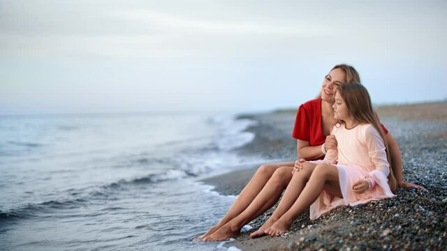 Happy elegant mother and daughter sitting on pebble beach coast together. Wide shot on RED camera
