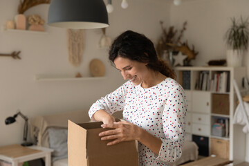 Plakat Excited millennial female open unpack postal cardboard box with internet order at home. Happy young woman unbox post package shopping online on web. Good delivery service, shipping concept.