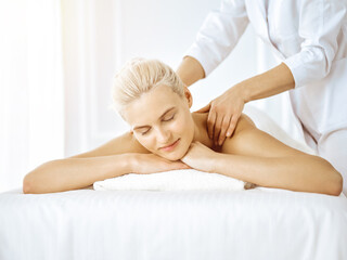 Obraz na płótnie Canvas Beautiful blonde woman enjoying back massage with closed eyes in sunny spa salon. Beauty concept