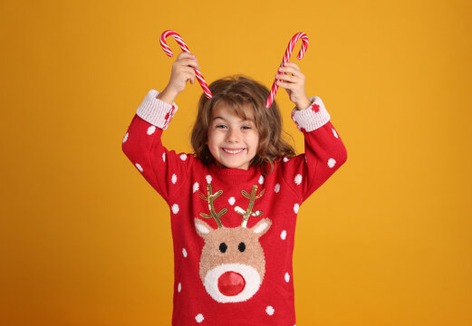 Cute Little Girl In Christmas Sweater Holding Sweet Candy Canes Near Head Against Orange Background