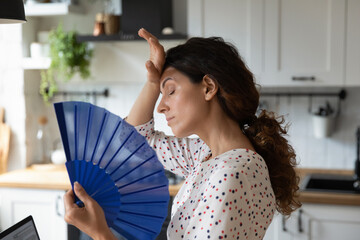 Unwell young Caucasian woman feel overheated at home wave with hand fan suffer from hot weather. Unhealthy tired female struggle with hormonal imbalance use waver. Air condition system.
