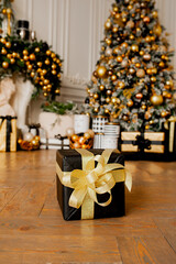 black gift box with gold ribbon on wooden floor in room with Christmas decor in gold and black colors : Christmas tree with balls and garlands and many Packed gifts under it, selective focus