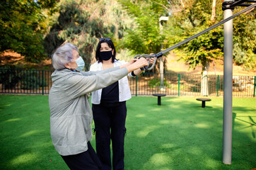 A physical therapist or professional caregiver watching attentively as a senior adult woman exercises on an outdoor fitness field. Women wear medical masks due to the pandemic.