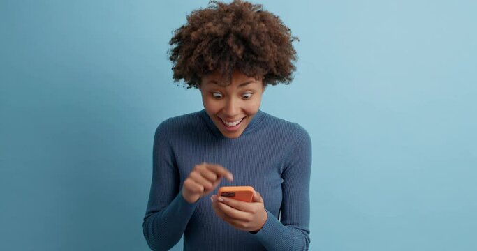 Young surprised excited young female model points at modern smartphone display reacts on unexpected discounts in online store wears casual turtleneck isolated over blue background waits for reply
