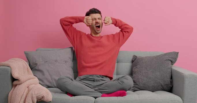 Sleepy young man yawns and stretches thinks what to do during leisure time sits crossed legs on comfortable sofa dressed in casual clothes isolated over pink background. Tired student indoor
