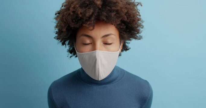 Health safety and coronavirus concept. Shocked curly woman stares bugged eyes at camera wears face protective mask tired of stress and tension wears casual turtleneck isolated over blue background