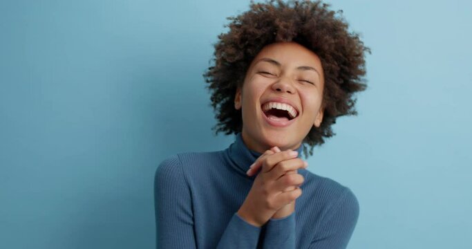 Overemotive dark skinned curly woman cannot stop laughing clasps hands and giggles positively hears positive story joke or funny anecdote dressed casually poses against blue background has fun