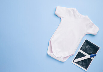 Ultrasound with a pregnancy test and clothes of a newborn baby on a blue background. Motherhood concept, white bodysuit for newborn. Space for text. 