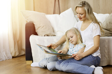 Happy family. Blonde young mother reading a book to her cute daughter while sitting at wooden floor in sunny room. Motherhood concept