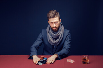 Stylish bearded Man in suit and scarf playing in dark casino, smoking cigar, drink whiskey.