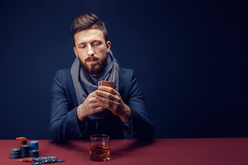 Stylish bearded Man in suit and scarf playing in dark casino, smoking cigar, drink whiskey.