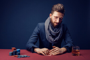 Stylish bearded Man in suit and scarf playing in dark casino, smoking cigar, drink whiskey.