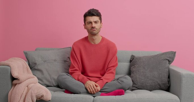 Serious unshaven man watches attentively football game makes face palm dissatisfed with results gestures with hands and asks why poses crossed legs on comfortable sofa against pink background