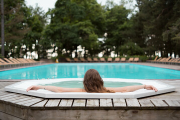 Photo of alone unrecognizable pretty girl sitting in warm water in outdoor pool. Photo from the back. Outside pool with turquoise water. Selective focus on girl. Outdoor recreation in spa hotel.