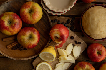 Ingredients of Apple Pie/Tarte Tatin on a wooden table: Dough, red apples, some sliced, one peeled, a lemon, raisins and pie dishes/tartelette forms. 