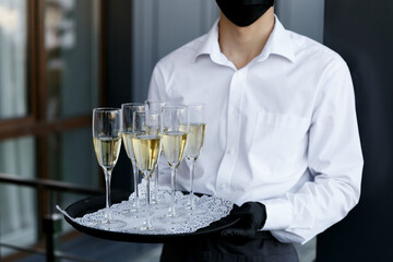 The waiter holds a tray with glasses of champagne. A waiter in a medical mask. Restaurant work during the coronavirus pandemic.