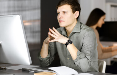 Young businessman and programmer in a green shirt is feeling stress after hard work, while sitting at the desk in a modern cabinet together with a female colleague at the background. Startup business