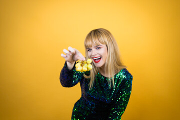 Beautiful blonde woman wearing a fancy dress over yellow background holding a bunch of grapes. New...