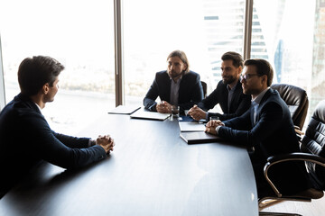 Businessmen sit at desk in boardroom talk discuss cooperation with client. Serious male recruiters employers have job interview with intern or work candidate at office meeting. Recruitment concept.