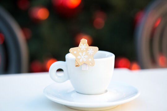 White porcelain coffee cup with saucer and wooden decorative star with snowflakes. Blurred background with chairs and Christmas tree with red balls. New year bar scene. Congratulations. Celebrating