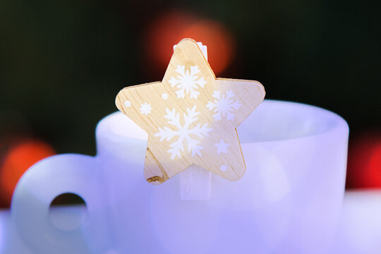 Wooden decorative star with snowflakes closeup with violet bokeh on white porcelain coffee cup. Blurred background with red Christmas lights. New year postcard. Congratulations.