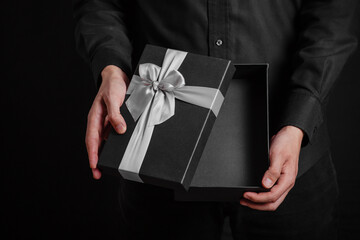 A guy in a black shirt on a black background holds an open gift box with a silver bow.