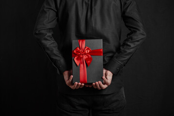 Man in a black shirt holds a gift with a red ribbon behind his back. Black gift box in hands. Surprise. Place for inscription.