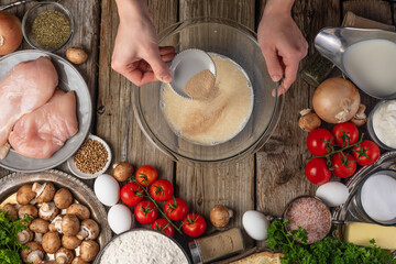 Chef hands pours spices into glass bowl with dough for pie on wooden table with variety of ingredients background. Concept of cooking process. Backstage of preparing tasty meal. View from above.