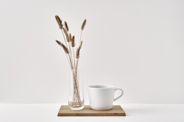 A vase with dry spikelets and a mug stand on a wooden stand on a white background. Minimalism, eco-materials in the interior decor. Copy space, mock up.