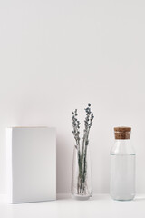 A transparent vase with lavender, a bottle of water with a cork stopper and a book on a white...
