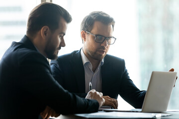 Focused Caucasian young male business partners or colleagues sit in office look at laptop screen brainstorm together. Serious businessmen work on computer talk discuss project. Cooperation concept.