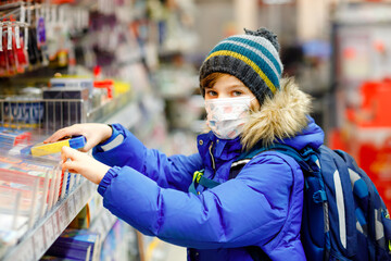 Little kid boy wearing medical mask buy school stuff in supermarket. Child with backpack and winter clothes. Schoolkid buying pencils. Lockdown and quarantine time during corona pandemic disease