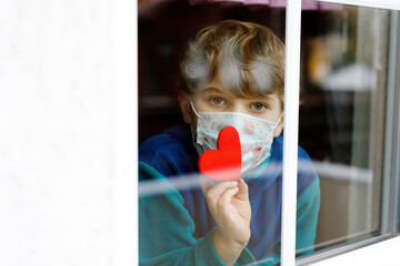 Lovely little school kid boy by a window wearing medical mask and holding wooden heart during...