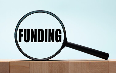 Cubes on a light blue wooden background. On them a magnifying glass with the word FUNDING