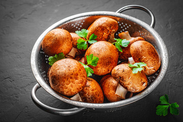 Fresh raw mushrooms champignons in stainless steel colander on Dark grey black slate background