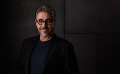 Obraz premium Portrait of happy older man in 50s with gray hair and glasses against dark background. Wearing jacket, smiling, copy space.