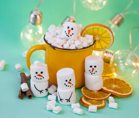 marshmallow snowmen on a mint turquoise background with a yellow cocoa mug illuminated by a garland