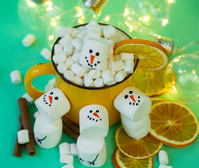 marshmallow snowmen on a mint turquoise background with a yellow cocoa mug illuminated by a garland