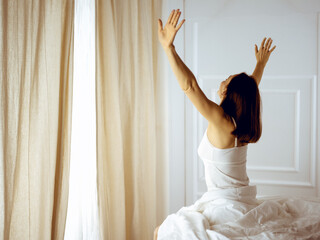 Woman stretching in bed after waking up, back view, entering a day happy and relaxed after good night sleep. Good morning and new day for brunette. Concept of Weekend, holidays