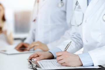 Unknown woman-doctors at work with patient at the background. Female physicians filling up medical documents or prescription while standing in sunny hospital reception desk, close-up. Health care