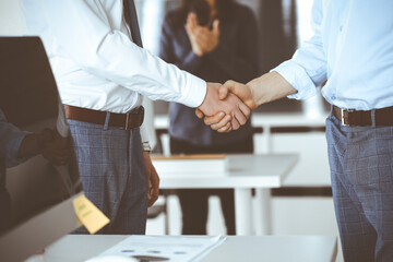 Fototapeta premium Two businessmen are shaking hands in office, close-up. Happy and excited business woman stands with raising hands at the background. Business people concept