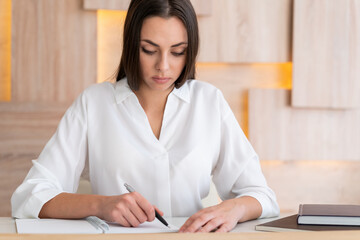 Office manager woman wearing white shirt, sitting to table with laptop and notebook, writing with a pen. Concentrated office worker in big light office room. Concept of work